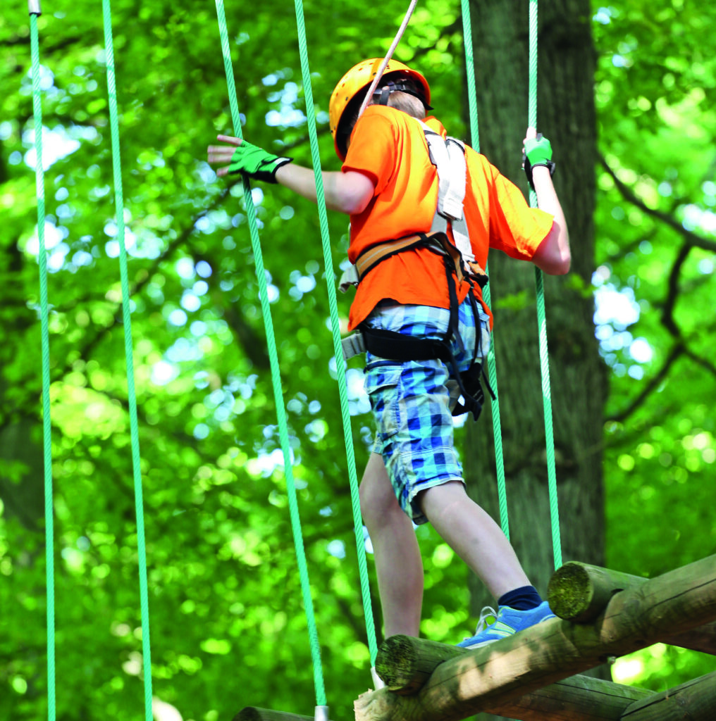 Eine Person balanciert auf einer Plattform im Kletterpark, umgeben von grüner Natur, bei sonnigem Wetter.