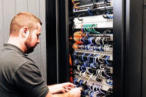 Eine Person arbeitet konzentriert an Kabeln in einem Server-Rack in einer IT-Umgebung.