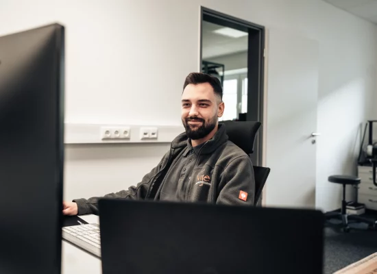 IT-Beratung-Vije Ein lächelnder Mann arbeitet entspannt an zwei Computern in einem hellen Büro.