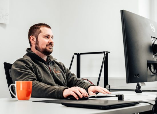 P1182315 Ein Mann arbeitet konzentriert am Computer in einem hellen, aufgeräumten Büro mit einer orangefarbenen Tasse.