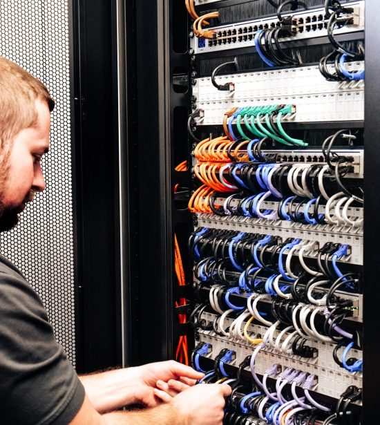 Eine Person arbeitet konzentriert an Kabeln in einem Server-Rack in einer IT-Umgebung.