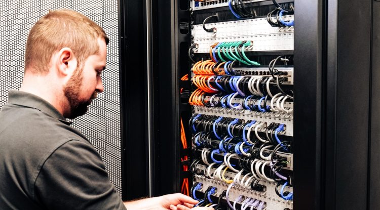 Eine Person arbeitet konzentriert an Kabeln in einem Server-Rack in einer IT-Umgebung.