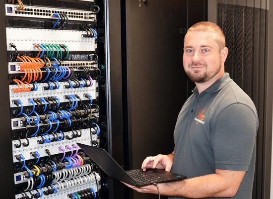 image Person im Serverraum arbeitet konzentriert am Laptop, umgeben von farbigen Kabeln und Servern.